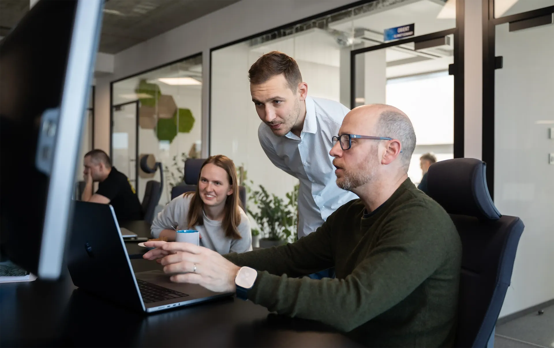 Three people in an office sit and stand around a laptop.